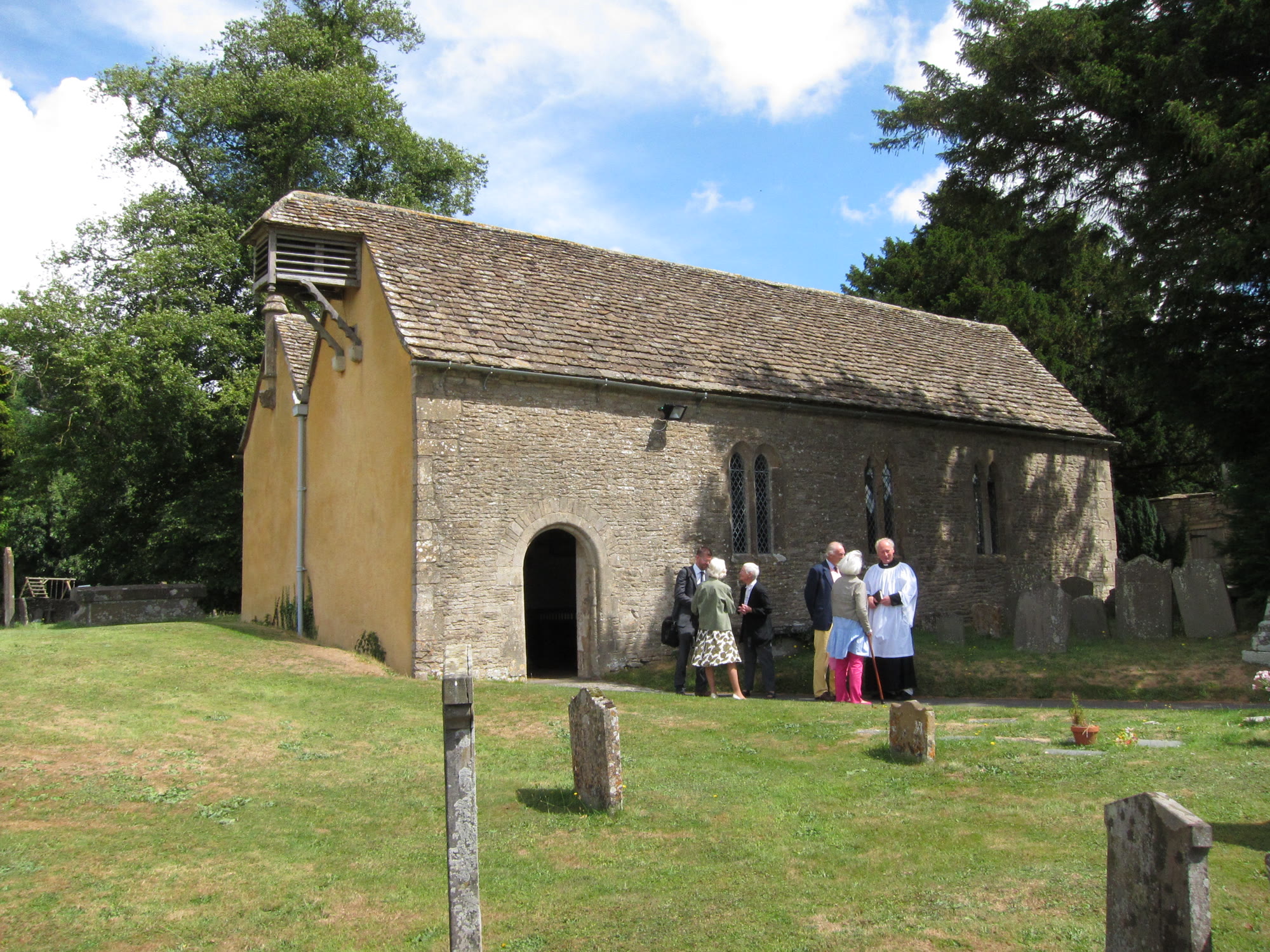 The village church at Little Badminton on a bright day, photographed by Adrian Pingstone on 28 July 2013. People in the churchyard are not identified.